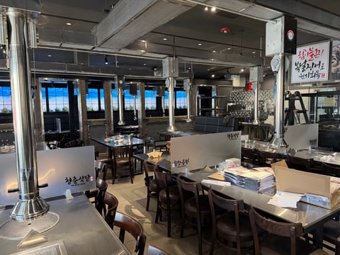 Modern restaurant interior with dining tables, wooden chairs, floor-to-ceiling windows overlooking a city skyline at dusk, and Korean signage on the walls