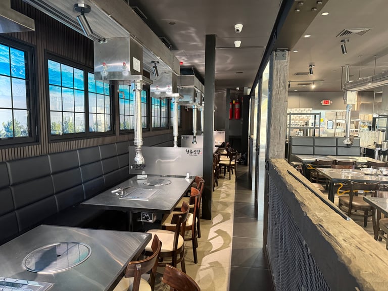 Modern restaurant interior with booth seating, exposed ceiling beams, large windows, and open kitchen view on the right side